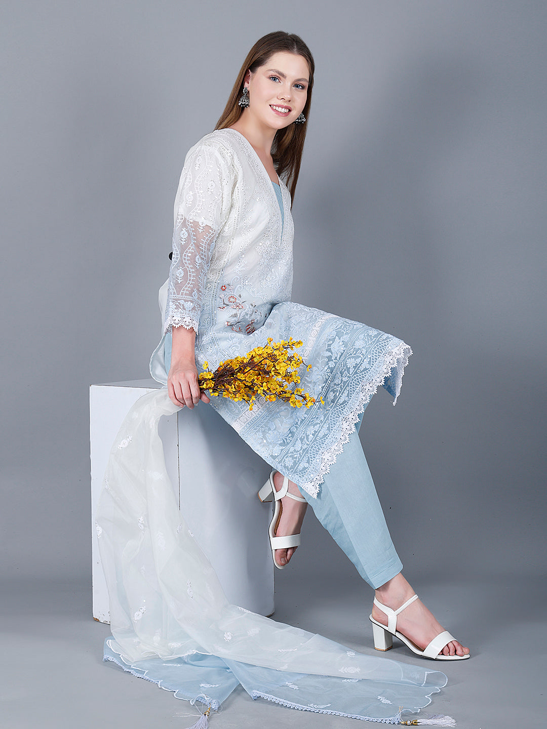 A woman sits on a white cube against a gray background, wearing the SUFIANA MEHAK RANGE P1 Pakistani Suit adorned with intricate embroidery. Holding a bunch of yellow flowers, she smiles warmly. Her long hair cascades down, and her white heels perfectly complement her traditional embroidered suit.